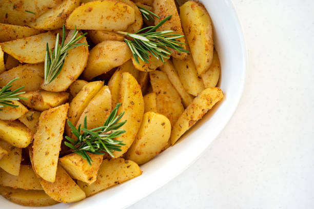 Roasted potato wedges with spicy herbs, spices and pieces of fresh rosemary on the white porcelain baking dish stock photo