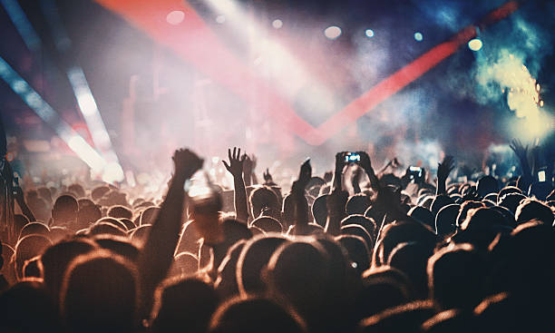 Rock concert. Rear view of people cheering at concert during music festival.Large group of unrecognizable adults with their arms raised.Some of them holding beer cans,some are taping the show with phones.There's a stage and a band performing in background,out of focus.Stage is toned purple blue with lasers and spotlights cutting through smoke and dust. dance music stock pictures, royalty-free photos & images
