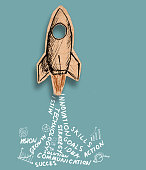 sketch of a rocket cut from brown crumpled paper with business words as engine smoke drawn on brown on a blue background