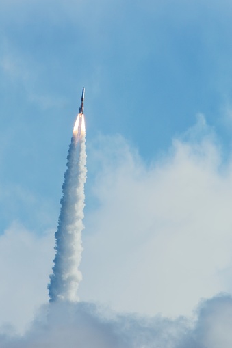 A Delta rocket passes through a cloud  propelled by a single main rocket engine and 2 booster rockets. Flames shoot from the booster rockets leaving behind a heavy exhaust plume.