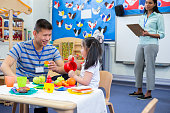 Male teacher playing in a toy kitchen with a nursery student. There is a teacher in the background with a clipboard who is watching them.
