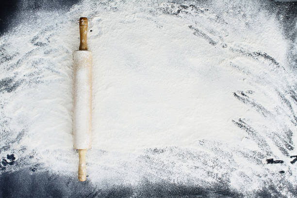 Rolling Pin Over Flour Background Old wooden rolling pin dusted with white flour over a flour covered dark background. Image shot from above. flour stock pictures, royalty-free photos & images