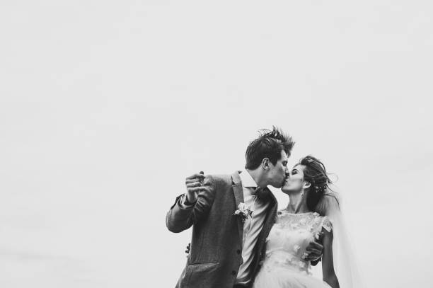 Romantic couple newlyweds, bride and groom is kissing on the background of a blue sky in park. Peaceful romantic and joyful wedding moment on nature in the field or water. Black and white photo. stock photo