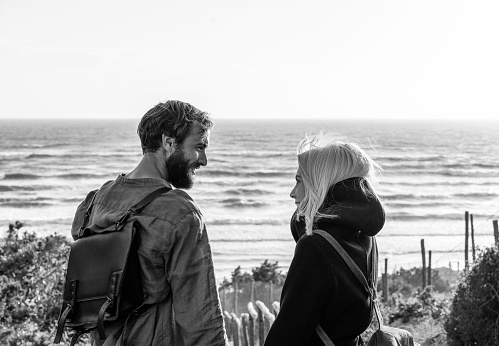 Romantic happy couple in love standing on wooden stairs on dunes at the wild beach in front of the sea - Boyfriend and girlfriend wearing vintage backpack looking into each other's eyes at sunset