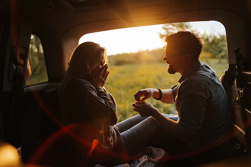 Romantic young couple sitting in open car trunk, enjoying sunset in spring nature while taking a break from long road trip, boyfriend using the romantic moment to propose his girl, she is happy and surprised