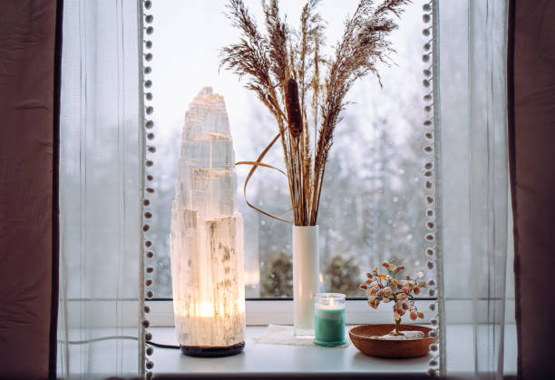 Rough big selenite crystal tower pole lamp illuminated on home window sill, spiritual home decor accent. Winter forest on background. stock photo
