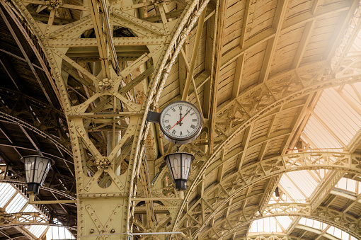 Round Roman clock and glass roof of railway or subway station. Twelve o'clock, seven past twelve pm, sunflare.