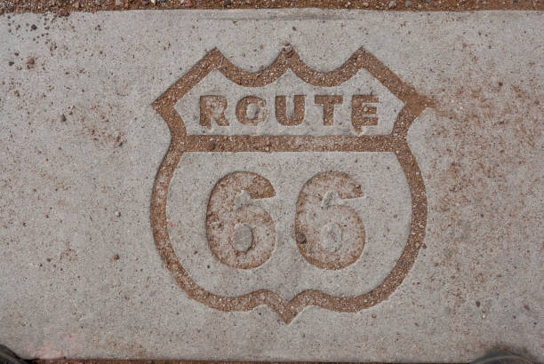 Route 66 Logo Engraved in Pavement Historic US Highway 66, known as the “Mother Road”, is more than just a stretch of pavement.  It is also an American icon, a symbol of opportunity, adventure and discovery.  US Highway 66 better known in literature, song, and story as Route 66, was a ribbon of roadway over two thousand miles long that connected Middle America to the Pacific coast.

Beginning in 1926, Route 66 fulfilled different needs in each subsequent decade.  In the 1930’s it was the main travel corridor for migrating families fleeing the Dust Bowl.  In the 1940’s, during World War II, the road was filled with military traffic.  In the 1950’s Route 66 came into its own as motorists took to the road to explore the nation with a freedom never felt before.  In the next decades as the interstate highway system was being developed, drivers bypassed Route 66 in favor of the faster freeways.  Unfortunately, the tourists also bypassed the small towns that gave the Mother Road its character and appeal.

Petrified Forest is the only National Park in the country that contains a section of Historic Route 66.  This stretch of Route 66 was open from 1926 until 1958 and was the primary way millions of travelers accessed the Petrified Forest and Painted Desert.  Near Tiponi Point and the Painted Desert a section of the original roadbed has been preserved along with interpretive signs and a rusty 1932 Studebaker.

The Route 66 logo is engraved in the pavement at the Route 66 interpretive center in Petrified Forest National Park near Holbrook, Arizona, USA. jeff goulden petrified forest national park stock pictures, royalty-free photos & images