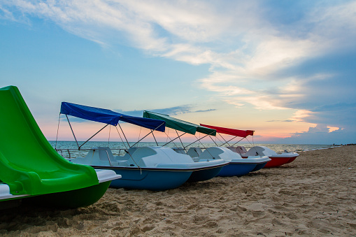 A row of colorful catamarans with canopies stand on the beach near the sea in the background of the setting sun