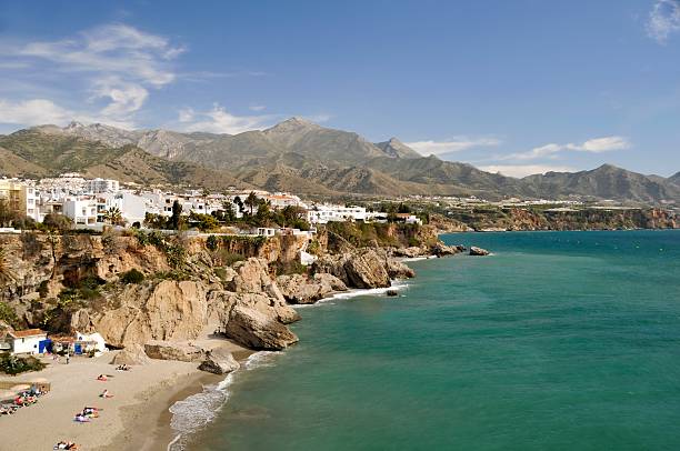 Rugged coastline in Nerja in Malaga, Spain The view of the Nerja in Malaga, Spain, is pictured.  There are mountains and an outcropping of rock to the left of the sea dotted with white and pale-colored houses.  The sea is teal and blue with a blue sky and wispy white clouds in the background. costa del sol málaga province stock pictures, royalty-free photos & images