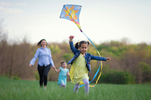 Sweet little girl running with a kite in front of his mother and sister. Female part of the family spending time in fun and games outdoor.
