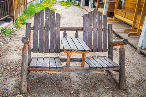 Rustic wooden seat for two with table built in middle of bench and arm rests and backs sitting on dirt in alleyway with porches with more wooden furniture to sides