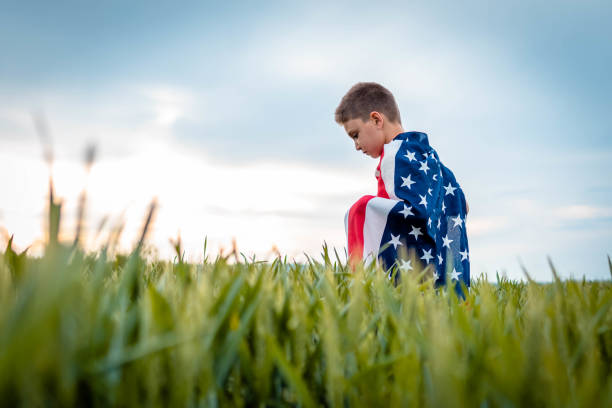 грустный мальчик, завернутый в американский флаг. он стоит на коленях на поле, глядя грустно - july 4 стоковые фото и изображения
