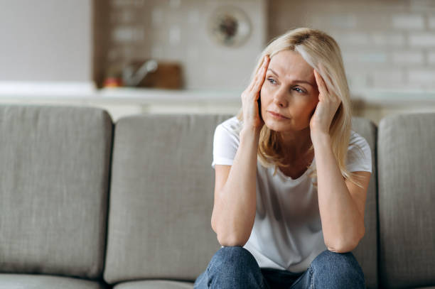Sad caucasian senior woman wearing casual clothes sits on couch at home alone feels unhappy because of headache, stress, illness or bad news, she needs rest and sleep stock photo