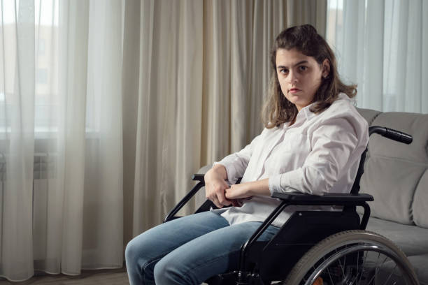 Sad disabled woman in wheelchair looks out of window at home stock photo
