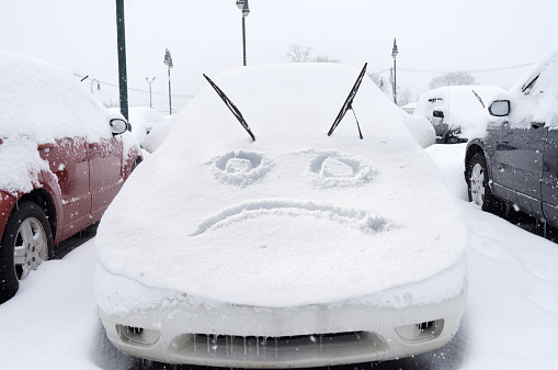 Winter storm car covered with snow with a sad face expression on hood of car. I hate winter theme. Winter sucks theme.