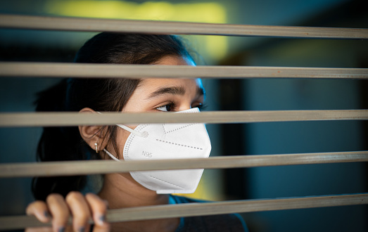 Indoor image of sad Asian/Indian teenager girl standing near house window with protective face mask and looking outside with a blank expression in lockdown for the covid-19 outbreak.