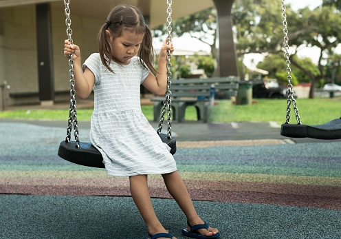 A little girl sitting on the swing lonely with no one to play with at the park playground.