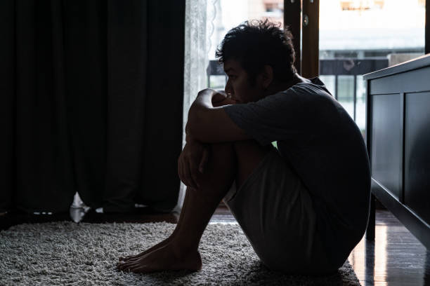Sad man sitting in bedroom Wide shot of young adult asian loneliness sad man sitting on the floor in bedroom hugging the knees and cry. Broken heart despair man sitting on the floor and thinking alone."n drug abuse stock pictures, royalty-free photos & images