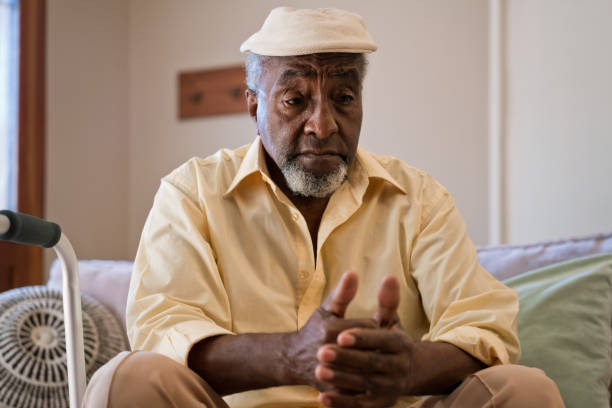 Sad senior man sitting in living room at home Depressed senior man in living room. Elderly male is looking down while sitting on sofa. He is wearing cap at home sad old black man stock pictures, royalty-free photos & images