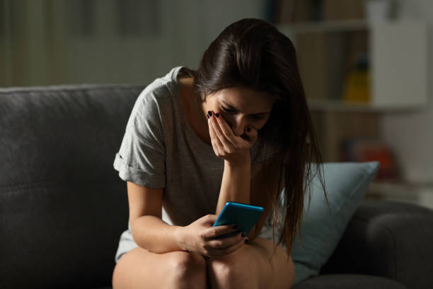 Sad teen receiving bad news online at home Sad teen receiving bad news online in a smart phone sitting on a couch in the living room at home anxiety photos stock pictures, royalty-free photos & images