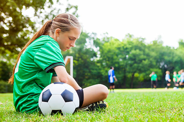 Sad teenage girl after soccer game Upset Caucasian female soccer player sits on the sidelines after losing the game. She is hanging her head down and is relective. She is wearing a green uniform and a soccer ball is beside her. Her teammates are on the field. She has blond hair in a pony tail. defeat stock pictures, royalty-free photos & images