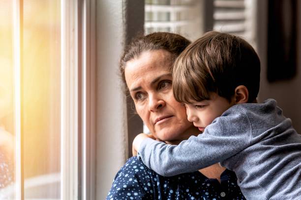 Sad Unemployed mature woman holding her son in her arms looking through a window stock photo