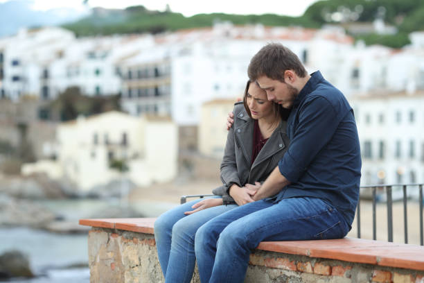 Sad woman and man comforting her on a ledge Sad woman and man comforting her on a ledge with a town in the background divorce beach stock pictures, royalty-free photos & images