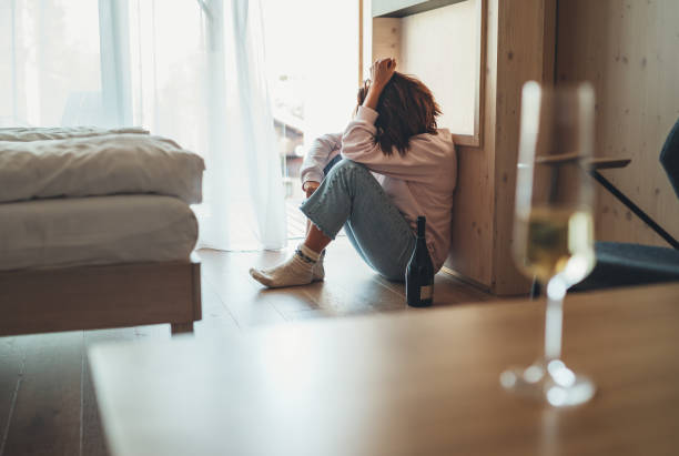 Sad woman sitting on the floor in the bedroom with a bottle of alcohol. Unfocused wine glass on the foreground table. Mental health and alcoholism problems concept image. stock photo