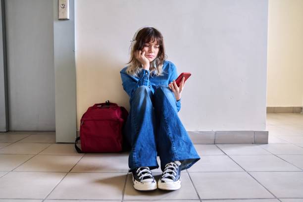 Sad young female student with backpack smartphone sitting on the floor stock photo