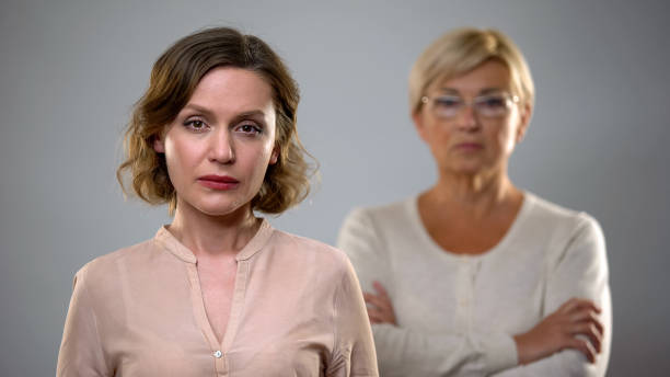 Sad young woman looking in camera, strict senior mother standing behind, problem stock photo