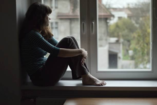 triest jonge vrouw zittend op het venster kijken - alleen één jonge vrouw stockfoto's en -beelden