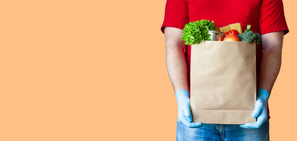 Safe food delivery concept Grocery delivery courier man in red uniform and medical gloves holds paper bag with food on orange background. Safe food delivery during quarantine, online shopping or donation concept. Copy space. food bank stock pictures, royalty-free photos & images