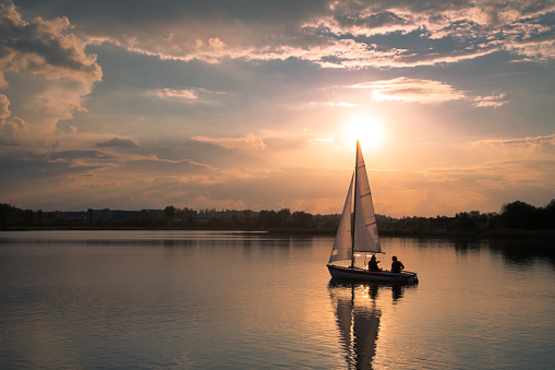 A couple sailing into the sunset. Yellow and orange sunlight, summer, calm sunny weather, clouds on the sky, small boat.