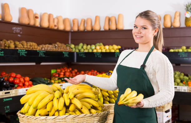 Salesgirl proposing bananas Positive female grocery worker in apron offering fresh ripe bananas salesgirl stock pictures, royalty-free photos & images