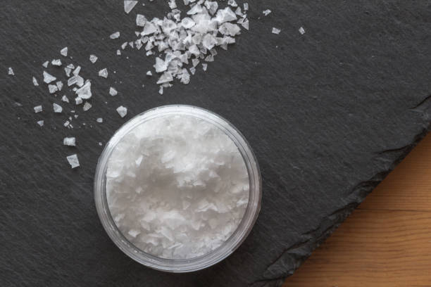Salt in a container with salt flakes on black slate background on wooden table stock photo