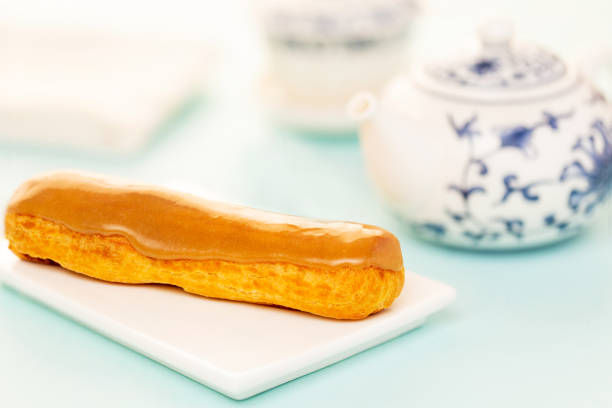 Salty caramel French eclair pastry on white plate, aside Chinese teapot over light blue background. stock photo