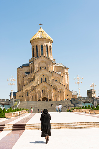 Tbilisi, Georgia - May 3, 2013: Sameba cathedral in Tbilisi. Long bright stone footpath leading to the cathedral, in front view a woman in black walking to the church, in distance some other people on the stairs in front of the church. Blue clear sky.