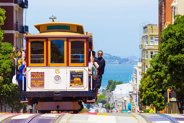 San Francisco Bay Coming Cable Car Front Close H San Francisco, United States - May 19, 2016: Approaching iconic cable car is full of outside hanging and platform standing tourists with bay water background in sunny Powell Street in California. Horizontal cable car stock pictures, royalty-free photos & images
