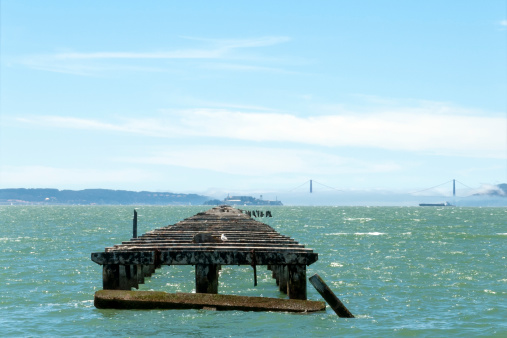 Remains of the Berkeley Pier, which once extended 3.5 miles into San Francisco Bay. The Golden Gate Bridge and Alcatraz are visible in the background, and a blimp is in the sky.
