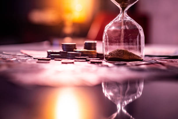 Sand running through the shape of hourglass on table with banknotes and coins of international currency. Time investment and retirement saving. Urgency countdown timer for business deadline concept stock photo