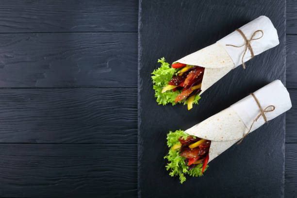 sandwich wraps with chicken meats and vegetables delicious freshly made sandwich wraps with frisee lettuce, sweet peppers, coleslaw and fried chicken sticks on black stone tray, view from above burger wrapped in paper stock pictures, royalty-free photos & images