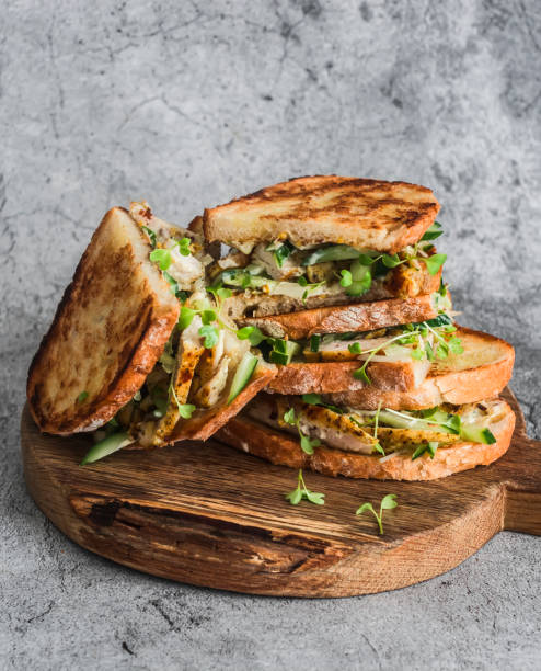 Sandwiches with turmeric fried chicken, cucumber, microgreens and homemade mustard mayonnaise sauce on a wooden cutting board on a gray background stock photo