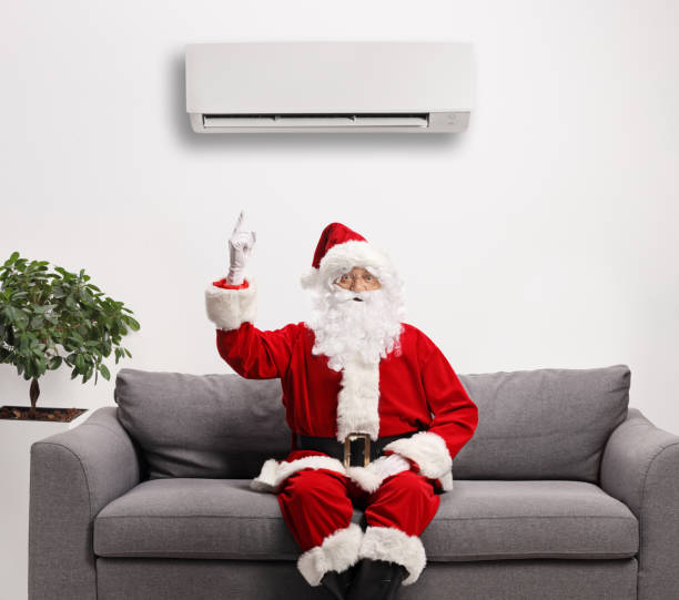 Santa claus sitting on a sofa and pointing to an air conditioning unit above stock photo