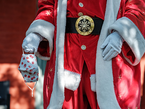 Santa's torso posing outdoors holding a protective  mask in his hand. Mask has a festive design to go with his jolly outfit.