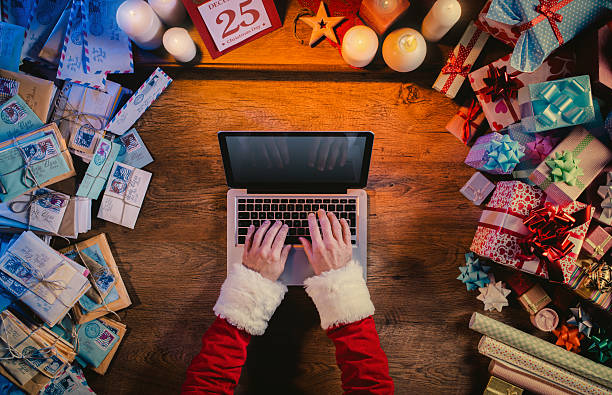 Santa working at desk and typing on a laptop surrounded by colorful...