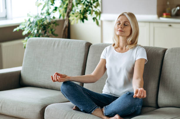 Satisfied attractive middle aged female sits on sofa at home and chilling. Blonde mature is practise yoga and meditation in lotus position with eyes closed, relieve stress, calmness concept stock photo