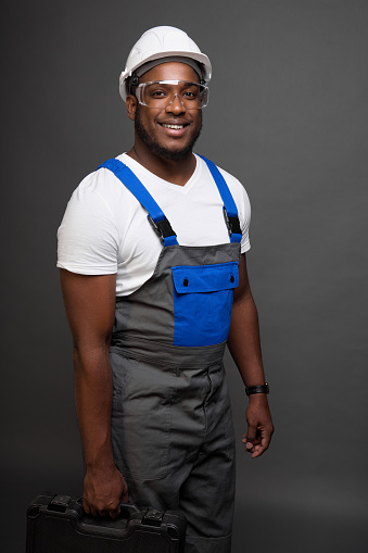 Smiling engineer in protective helmet and transparent glasses stands against gray wall holding suitcase with tools in hand. Satisfied black engineer with box professional tools in his hand looks ahead