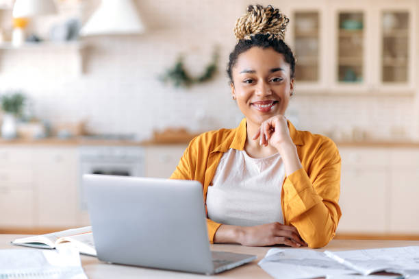 Satisfied good looking young African American stylish woman, freelancer, student or real estate agent, sitting at her desk at home office, looking at the camera and smiling pleasantly stock photo