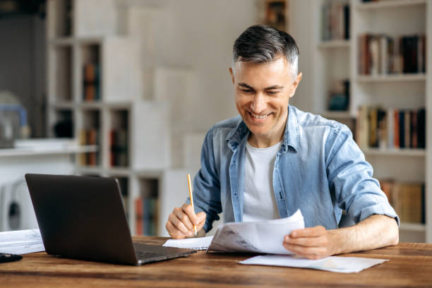 Satisfied successful confident mature gray-haired caucasian businessman, manager or freelancer, in casual stylish wear, working at office using laptop, checking work papers, taking notes, smiling stock photo